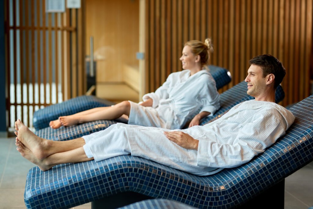 Smiling couple in bathrobes relaxing on lounge chairs at the spa